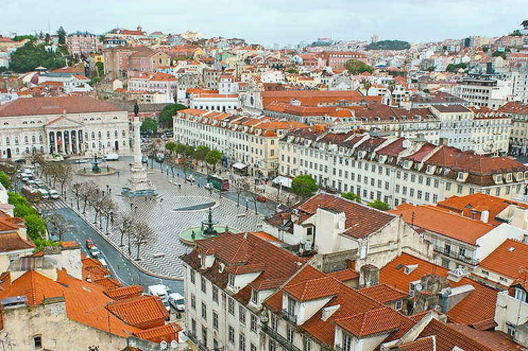 Imagem aérea da Praça do Rossio em Lisboa, mostrando edifícios com telhados vermelhos e uma ampla praça central. O céu está nublado e a cidade se estende ao fundo, com várias construções e ruas visíveis