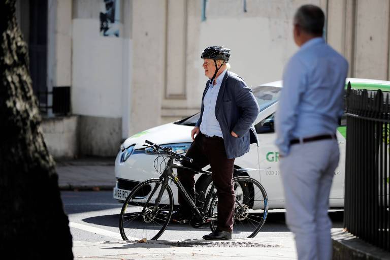 Johnson usa terno sem gravata e capacete. Ele está montado na bicicleta, de cor preta, apoiando na calçada com o pé esquerdo. A seu lado há um carro branco e verde. Um homem fala ao celular à frente da esquina.