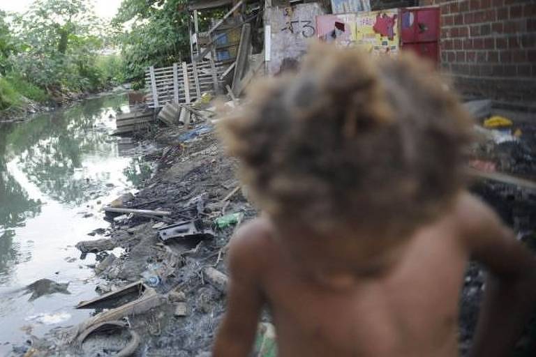 Criança ao lado de valão a céu aberto no Complexo da Maré, no Rio de Janeiro; violação do saneamento é quesito que afeta o maior número de meninos e meninas no país