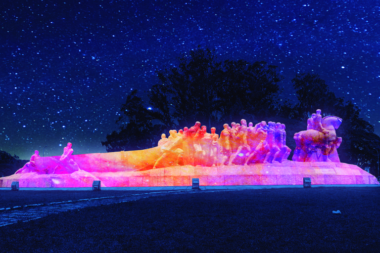 Simulação do monumento às Bandeiras iluminado, à noite, por projeção cor de rosa e laranja