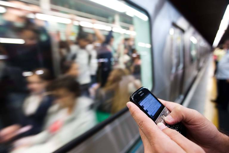Detalhe das mãos de uma pessoa, com telefone celular, em frente a um vagão do metrô em movimento
