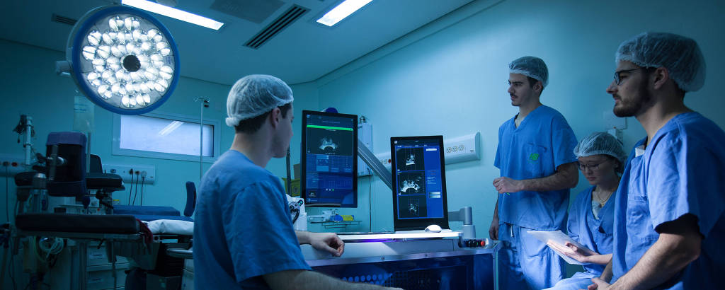 Grupo de três homens e uma mulher paramentado com roupas azuis e toucas para cirurgia diante aparelhos eletrônicos com dados vitais