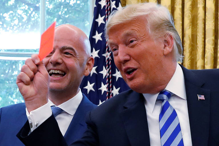 A imagem mostra Donald Trump， à direita， segurando um cartão vermelho e sorrindo， enquanto Gianni Infantino， à esquerda， também sorri. Ao fundo， há uma cortina dourada e a bandeira dos Estados Unidos.
