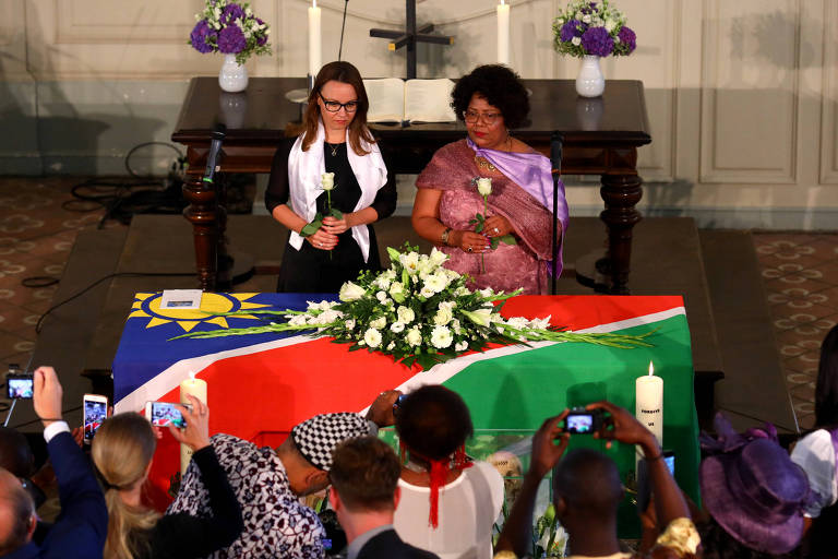 Duas mulheres velam caixão sob bandeira da Namíbia e grupo de pessoas acompanha a cerimônia