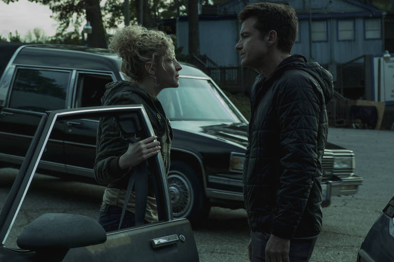 A atriz Julia Garner segura a porta de um carro no estacionamento de uma pousada ao cair de uma tarde nublada enquanto discute, séria, com o ator Jason Batemen, que a encara, em cena de Ozark
