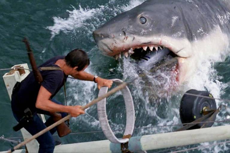 Um homem em um barco está tentando acertar um arpão dentro da boca de um grande tubarão que emerge da água. O tubarão tem a boca aberta， mostrando dentes afiados， enquanto o homem e parece estar em ação para capturá-lo. O cenário é agitado， com ondas e respingos de água ao redor.