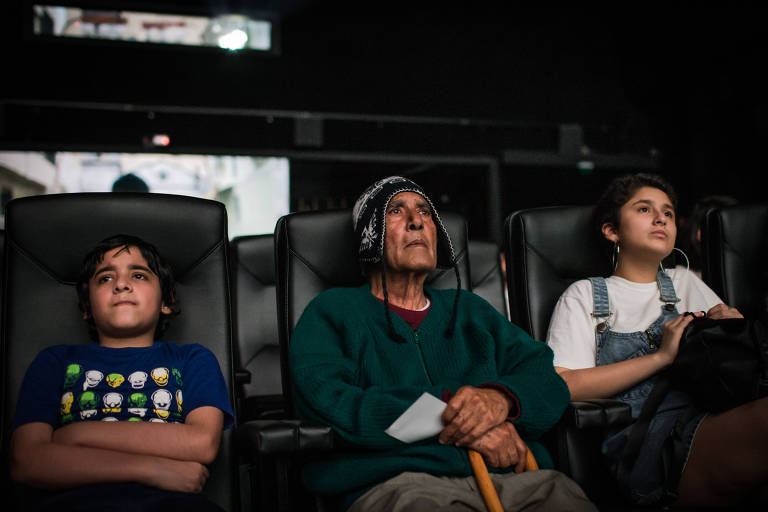 Três pessoas assistem filme em sala de cinema. Da esquerda para a direita: um garoto com os braços cruzados, um senhor com gorro andino e bengala e uma adolescente de camiseta branca e macacão