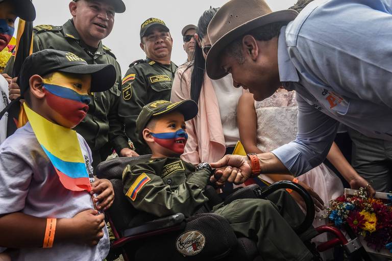 De camisa jeans clara e chapéu marrom, Duque se agacha para cumprimentar a criança, que está com o rosto pintado com as cores da bandeira da Colômbia. Ao lado do menino há outro também com o rosto pintado e ao fundo aparecem dois policiais e outras duas pessoas de pé.