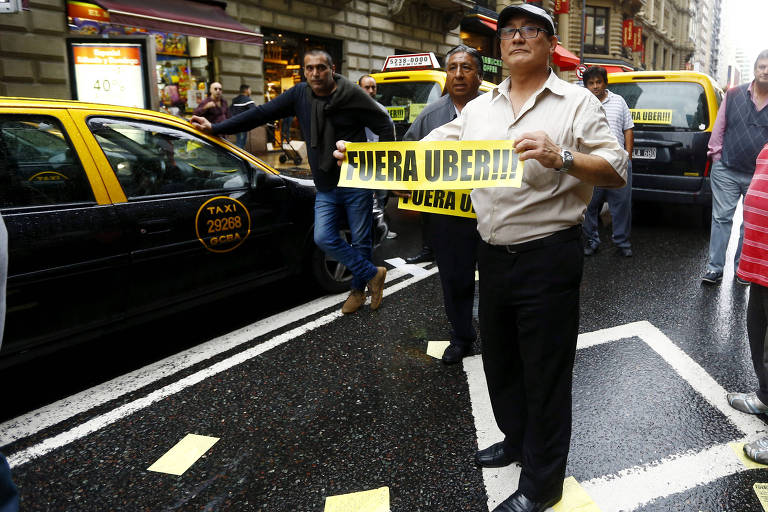 Homem carrega faixa com frase "Fora, Uber" ao lado de outros colegas e de fila de táxis parados em avenida