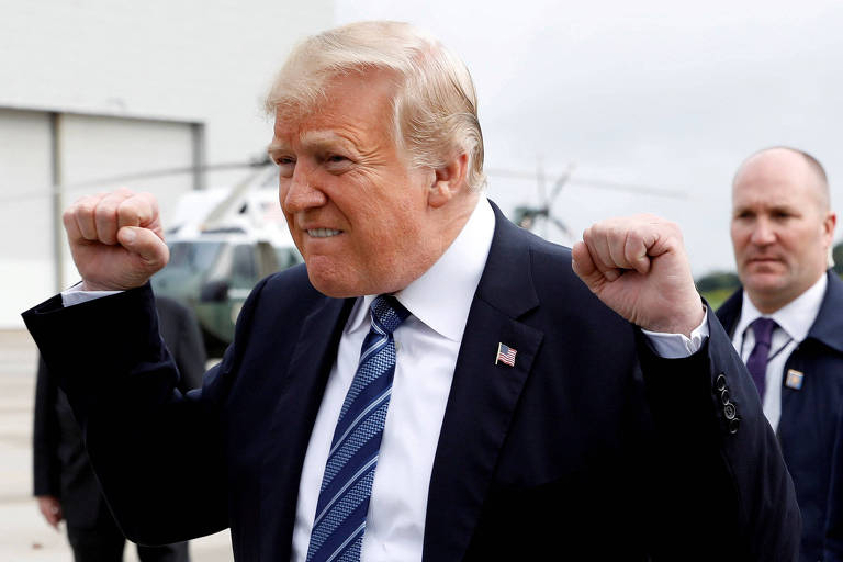 Trump franze o lábio e levanta as duas mãos fechadas. Ele usa terno preto, camisa branca e gravata azul. Atrás dele aparecem dois agentes do Serviço Secreto.