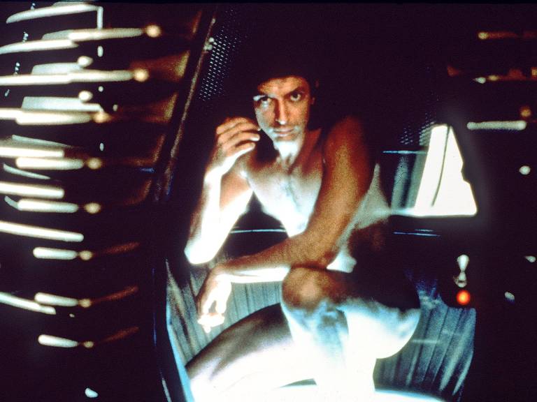 Goldblum, um homem moreno, está agachado, nu, dentro de uma cápsula metálica. Ele olha diretamente para a câmera