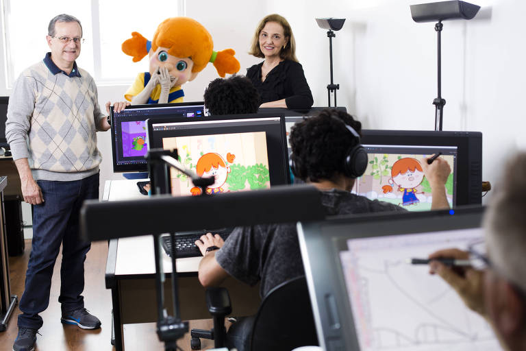 Dario e Elizabeth com uma pessoa vestida como uma personagem de cabelos laranjas e maria chiquinha, dentro do estúdio da empresa com pessoas trabalhando no computador e desenhando