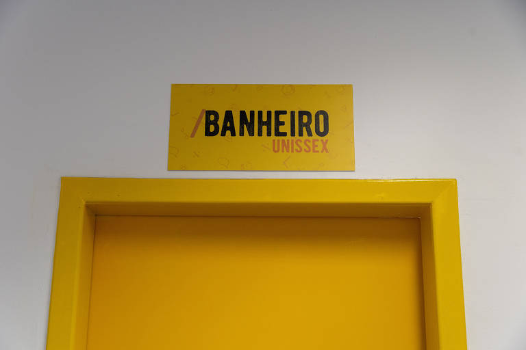 Imagem mostra porta amarela de banheiro unissex. Há uma placa indicando que o sanitário não distingue gêneros.
