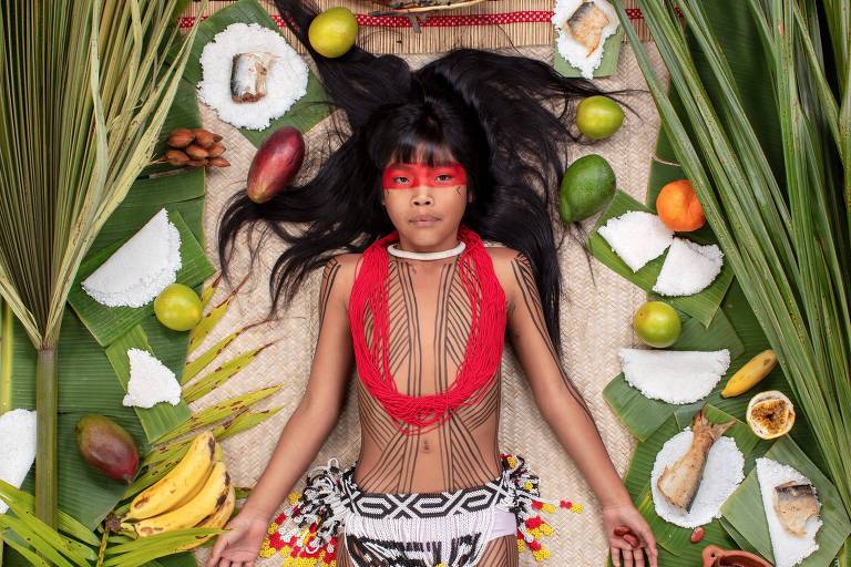 Imagem mostra criança indígena com pintura vermelha sobre os olhos e ao seu lado há alimentos como banana e tapioca
