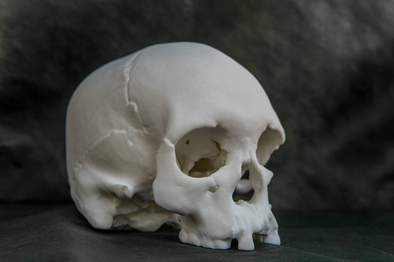 Imagem em 3D de um crânio branco de cor branca, levemente virado para direita, em cima de um tecido preto, com um fundo de tecido preto atrás