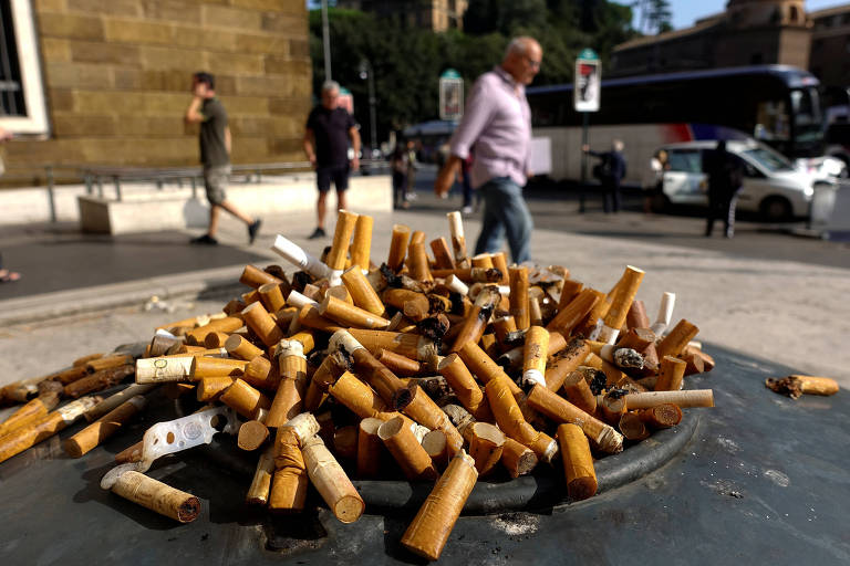 Montes de cigarros estão despejados em um cinzeiro colocado em cima de uma mesa; ao fundo， um homem caminha