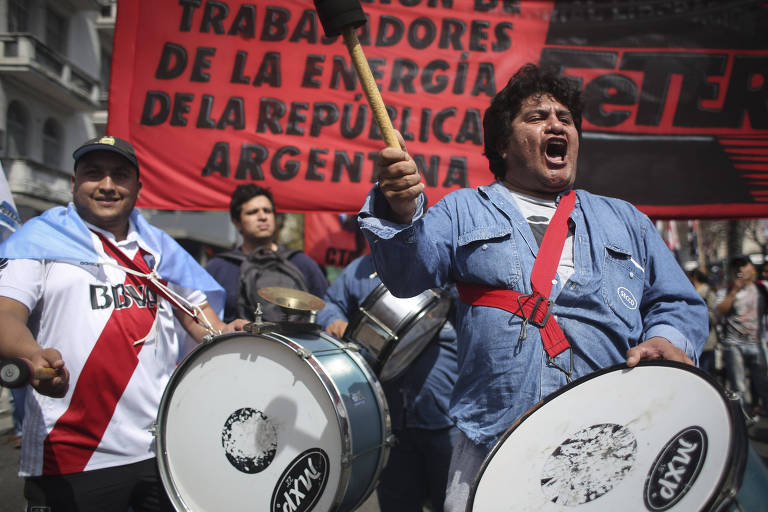 Dois sindicalistas aparecem tocando tambores. Atrás, há uma faixa vermelha com letras pretas do sindicato dos trabalhadores da energia da República Argentina