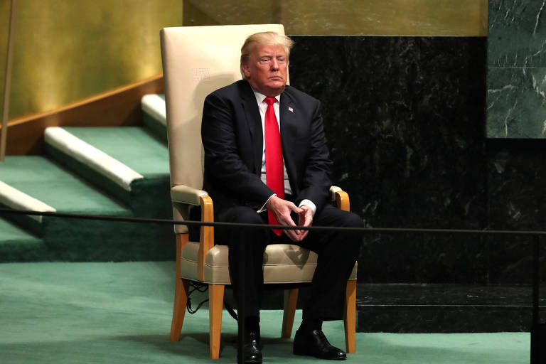 Trump está sentado em uma poltrona de madeira, com encosto de cor bege, no palco da Assembleia-Geral da ONU, que tem um carpete verde. Ao fundo, aparece parte da mesa diretora, de cor preta.