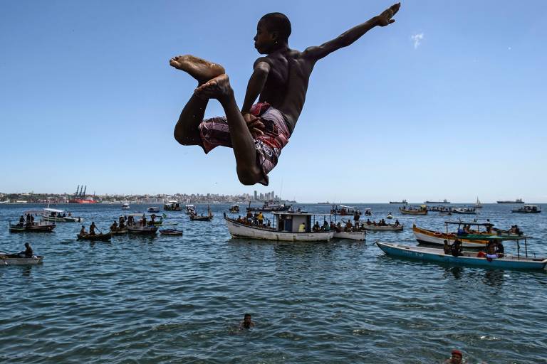 Menino negro, usando short vermelho e branco, salta para pular no mar, em uma praia de Salvador. Na água, é possível ver dezenas de barcos.