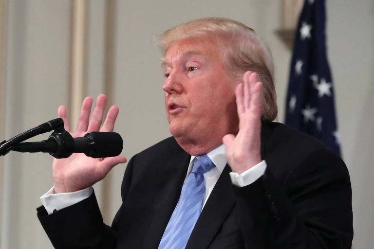 Trump faz cara de espanto e levanta as duas mãos enquanto fala ao microfone. Ele, que usa terno preto, camisa branca e gravata azul clara, aparece de perfil. Ao fundo, uma bandeira americana.