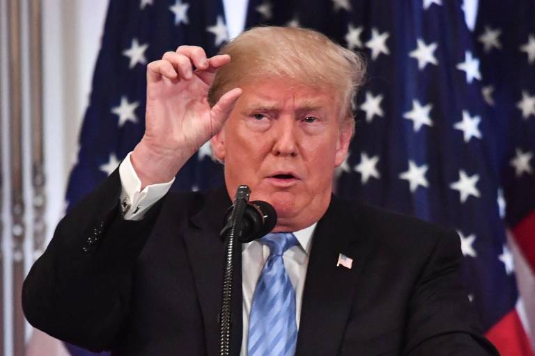 Trump faz sinal com os dedos polegar e indicador da mão direita. Ele, que usa terno preto, gravata azul e camisa branca, fala em um púlpito. Atrás, aparecem duas bandeiras americanas.