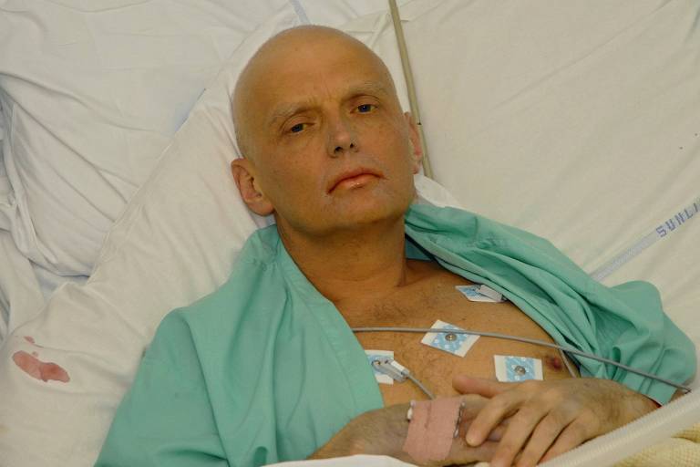 Litvinenko está em uma cama de hospital, usando um pijama verde aberto, com eletrodos no peito