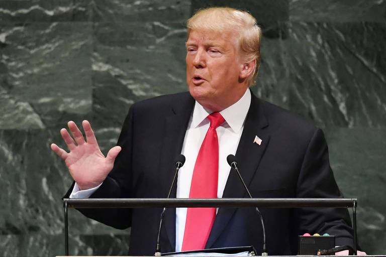 Trump levanta a palma da mão esquerda enquanto fala. Ele usa um terno preto, camisa branca e gravata vermelha. Ao fundo, uma parede de pedras verdes.