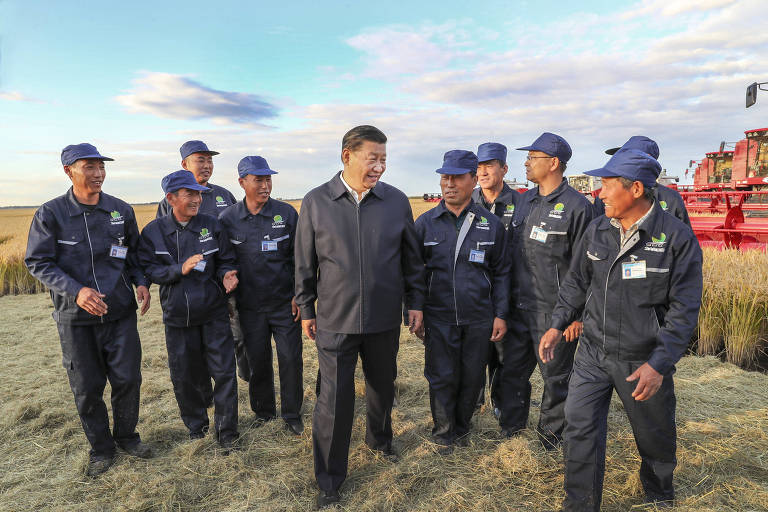 Xi Jinping aparece no meio da foto, com quatro funcionários uniformizados do lado esquerdo e cinco do lado direito. Ao fundo, uma área aberta de campo com uma colheitadeira vermelha.