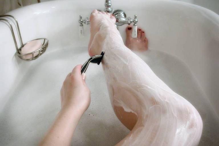 Imagem mostra perna de uma mulher dentro da banheira. A perna está coberta por creme branco e ela passa a lâmina de depilar com a mão esquerda