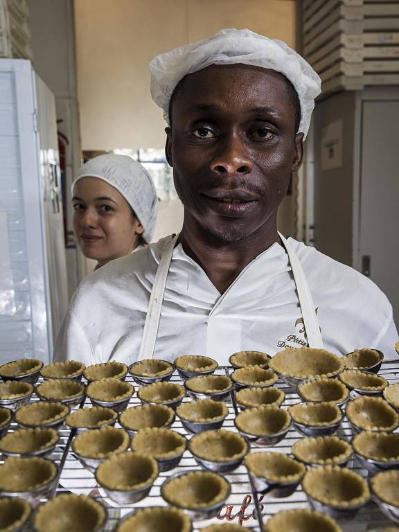 Retrato do confeiteiro haitiano Gabriel Lesli, 37, ao lado da confeiteira brasileira Bruna Britto (ao fundo), na cozinha da Patisserie Douce France. Ele trabalha no local desde junho de 2015 e foi um dos beneficiados pela regularização dos haitianos pelo Governo Brasileiro