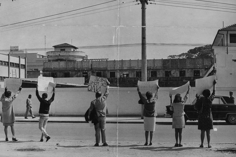 Foto em preto e branco de pessoas segurando cartazes no lado de fora de uma prisão