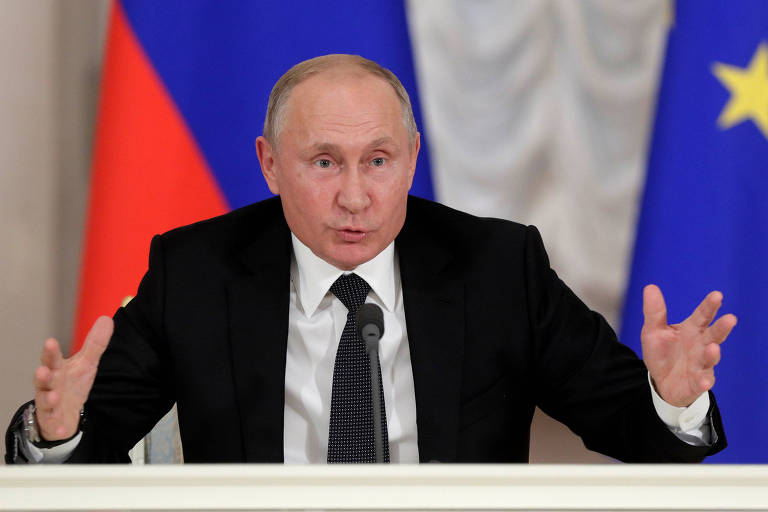 Putin, que usa terno preto, camisa branca e gravata preta, abre as mãos enquanto fala sentado à uma mesa. Ao fundo, bandeiras da Rússia. 