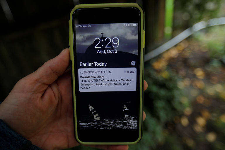 Mão de homem aparece com um celular preto com uma capa amarela fluorescente com o alerta na tela inicial.