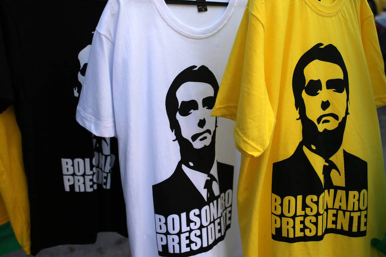 Três camisetas com fotos de Bolsonaro são vistas em uma arara. Da esquerda para a direita, uma preta, uma branca e uma amarela.