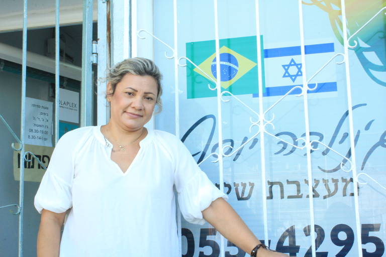 Genilda José aparece na frente do salão, que tem letreiro em hebraico com as bandeiras do Brasil e de Jerusalém