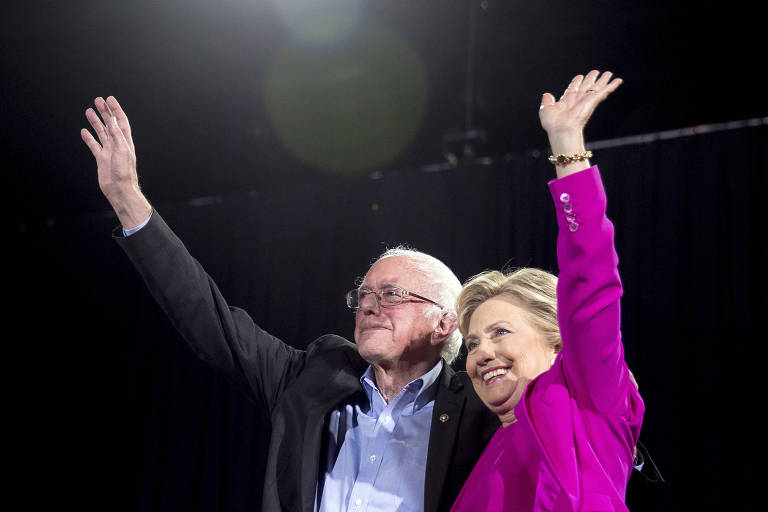 Hillary, de terninho rosa, está à direita, enquanto Sanders aparece à direita de paletó preto e camisa azul. Os dois levantam as mãos.