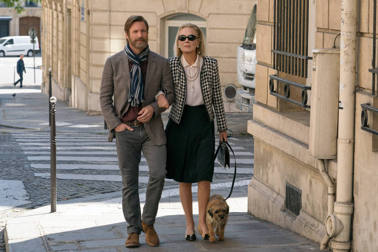 O ator Aaron Eckhart e a atriz Marthe Keller passeiam de braços dados, ela segurando um cachorrinho e usando óculos escuros, por rua em Paris 