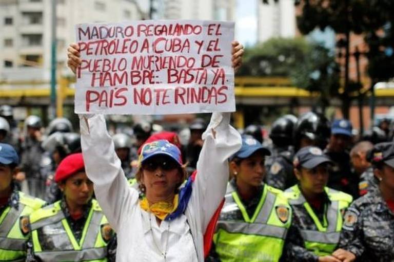 Manifestante carrega cartaz com a mensagem "Maduro dando petróleo de presente a Cuba e o povo morrendo de fome. Chega. País, não se renda". Ao fundo policiais fazem contenção.