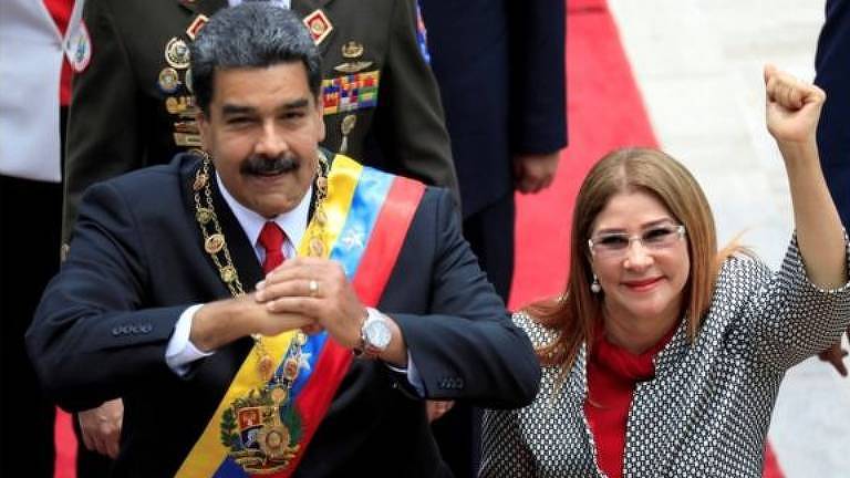 Maduro faz sinal com o punho de uma mão na palma da mão da outra, enquanto sua mulher, Cilia Flores, aparece à direita com o braço levantado