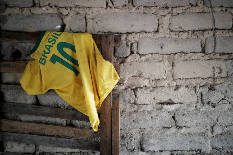 Camiseta aparece pendurada pelas costas em um estrado de madeira encostado em uma parede de tijolos cinza. Sua parte de trás tem a palavra Brasil e o número dez.