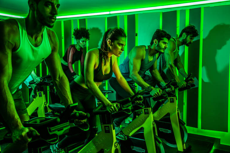 Em um salão iluminado de verde, pessoas correm em bicicletas dentro da academia 