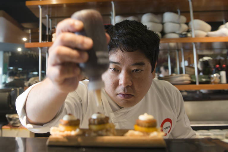 Em uma foto retangular se vê a mão do chef Mitsuharu "Micha" Tsumura apertando uma bisnaga para finalizar um prato; seu restaurante Maido, fica no Peru