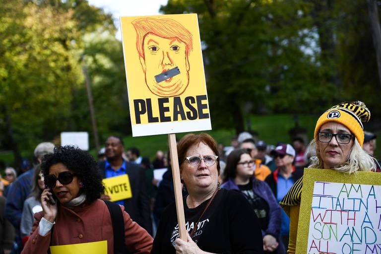 Três mulheres aparecem à frente do protesto, segurando cartazes. A do meio segura uma placa alta amarela, com uma caricatura do presidente com um adesivo preto na boca e a expressão por favor