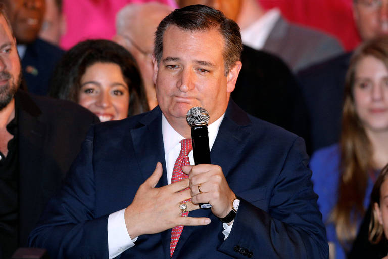 Ted Cruz segura um microfone com a mão direita, enquanto põe a mão esquerda no peito. Ao fundo, um grupo de apoiadores o observa.