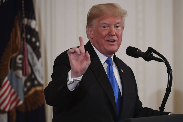 Vestindo terno preto com gravata azul, Trump aparece de perfil, falando em um púlpito com microfones. Ele levanta o dedo indicador da mão direita enquanto fala.