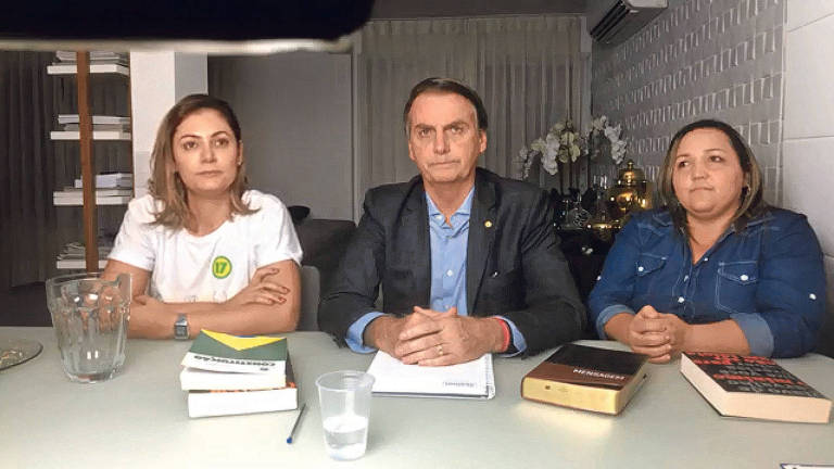 O presidente eleito aparece em vídeos improvisados e com estética que reforçam apelo popular; no vídeo em que Bolsonaro faz discurso após vitória, aparece ao lado da esposa Michelle (esq) e de uma tradutora de libras, na mesa podem ser vistos um copo plástico com água, uma jarra de vidro vazia e um prato com farelos