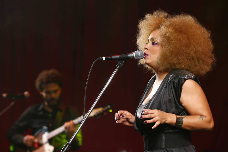 De vestido preto, cantora Anelis Assumpção canta ao microfone; ao fundo, à esquerda, Saulo Duarte toca guitarra
