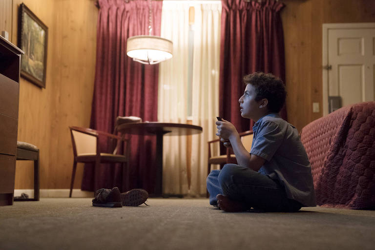 O ator adolescenteestá sentado no chão de um quarto de hotel acarpetado assistindo a TV