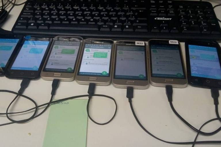 Sete celulares estão em cima de uma mesa branca, conectados aos fios do carregador. Atrás dos aparelhos, há o teclado de um notebook.