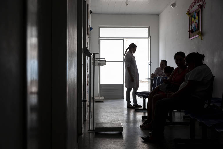 Pacientes em sala de espera; em pé, uma mulher com jaleco é vista contra a luz